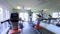 This fitness center offers a clean, functional workout space designed to support a variety of cardio-focused exercise routines. The room is finished with light green walls and rubber flooring, providing a practical and durable surface suited for high-traffic use. Large windows along the back wall allow ample natural light to fill the space, contributing to an open and energizing atmosphere. A ceiling fan overhead aids in air circulation during workouts, complementing the overhead lighting fixtures. The cardio equipment selection is well-rounded, featuring treadmills, an elliptical cross-trainer, and a stationary bike, giving users multiple options for cardiovascular training. A wall-mounted television is positioned near the windows, allowing users to stay entertained during longer sessions. Full-length mirrors line one wall, visually expanding the space and allowing users to monitor their form. The combination of natural light, mirrored walls, and a thoughtfully arranged equipment layout makes this fitness center a comfortable and efficient space for daily exercise.