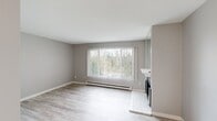 This single-floor unit offers a thoughtfully organized layout comprising a bedroom, bathroom, kitchen, and hallway. The interior is finished with light wood-style laminate flooring throughout, complemented by neutral gray walls and crisp white trim, creating a clean and cohesive aesthetic.

The living area is anchored by a large window fitted with vertical blinds, framing outdoor views of surrounding greenery and allowing ample natural light to fill the space. A baseboard heating unit runs beneath the window, and a tiled fireplace with a raised hearth serves as a functional focal point along one wall.

The galley-style kitchen is efficiently designed with gray shaker-style cabinetry, light quartz countertops, an undermount sink, and a full suite of appliances including an electric range and refrigerator. Track lighting above enhances visibility in the space.

The bedroom is a comfortable size, featuring a ceiling fan with light fixture, a window with vertical blinds, a baseboard heater, and mirrored sliding closet doors that reflect light and add a sense of openness.

The bathroom includes a full tub-shower combination with a white surround, brushed nickel fixtures and grab bar, a white vanity with countertop sink, and wood-style flooring that ties the space together with the rest of the unit.