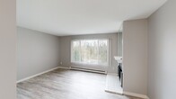 This single-floor unit offers a thoughtfully organized layout comprising a bedroom, bathroom, kitchen, and hallway. The interior is finished with light wood-style laminate flooring throughout, complemented by neutral gray walls and crisp white trim, creating a clean and cohesive aesthetic.

The living area is anchored by a large window fitted with vertical blinds, framing outdoor views of surrounding greenery and allowing ample natural light to fill the space. A baseboard heating unit runs beneath the window, and a tiled fireplace with a raised hearth serves as a functional focal point along one wall.

The galley-style kitchen is efficiently designed with gray shaker-style cabinetry, light quartz countertops, an undermount sink, and a full suite of appliances including an electric range and refrigerator. Track lighting above enhances visibility in the space.

The bedroom is a comfortable size, featuring a ceiling fan with light fixture, a window with vertical blinds, a baseboard heater, and mirrored sliding closet doors that reflect light and add a sense of openness.

The bathroom includes a full tub-shower combination with a white surround, brushed nickel fixtures and grab bar, a white vanity with countertop sink, and wood-style flooring that ties the space together with the rest of the unit.