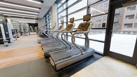 Located at 1213 Walnut St in the heart of Philadelphia, this modern fitness center offers a well-appointed, single-floor workout facility designed to meet the needs of serious fitness enthusiasts. The space is defined by its striking floor-to-ceiling glass window wall, which floods the interior with abundant natural light and frames dynamic views of the surrounding urban streetscape. The open layout creates a spacious, airy atmosphere that feels both motivating and comfortable. A row of commercial-grade Star Trac treadmills lines the window wall, allowing users to exercise while taking in the city views — a standout feature of the facility. The flooring transitions between durable rubber matting in the equipment zones and hardwood-style surfaces, providing both functionality and visual warmth. The high ceilings, contemporary architectural finishes, and clean, minimalist design contribute to a professional gym environment. Additional cardio and strength equipment is visible throughout the space, suggesting a comprehensive offering for a full-body workout. The overall ambiance is sleek and modern, combining industrial design elements with an inviting openness that makes it a premium fitness destination in Center City Philadelphia.