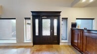 This single-floor facility spans a generously sized footprint, thoughtfully designed to accommodate both client-facing and active-use areas. The entrance makes a strong first impression with a bold black front door featuring diamond-patterned leaded glass and flanking sidelights, set beneath a tall, soaring ceiling that fills the foyer with a sense of openness and grandeur. Travertine tile flooring runs throughout the entry and reception zones, lending warmth and a polished, professional aesthetic.

Immediately inside, a substantial dark-wood reception desk anchors the front-of-house area, positioned to welcome visitors as they enter. Adjacent to the desk, a comfortable lounge area features upholstered seating, decorative greenery, and recessed lighting, creating an inviting atmosphere for waiting clients. High transom windows allow natural light to filter into the upper reaches of the lobby.

Beyond the reception area, the space transitions into an expansive fitness center divided into multiple exercise zones. Rubber flooring provides a durable, cushioned surface throughout. Equipment includes treadmills, ellipticals, stationary bikes, and free weights. Full-length mirrors line one wall, enhancing the sense of depth. Ceiling fans, wall-mounted televisions, and direct exterior access through glass doors complete this well-appointed fitness environment.
