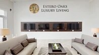 The Estero Oaks Luxury Living clubhouse at 9940 Estero Oaks Dr in Fort Myers, Florida, is an expansive, single-floor community facility that exudes a refined, contemporary elegance throughout. The space is finished with large-format light stone tile flooring that runs continuously from the grand entrance hallway through the main living and lounge areas, lending a cohesive and polished aesthetic. The welcoming hallway features a symmetrical layout with accent chairs, a decorative round mirror, and glass entry doors that frame lush views of swaying palm trees outside. Soaring ceilings — rising dramatically in key areas — are adorned with ornate chandeliers and intricate decorative grillwork, amplifying the sense of grandeur. Floor-to-ceiling windows bathe the main lounge in abundant natural light while offering unobstructed views of the outdoor pool area and pergola beyond. The living room is thoughtfully appointed with multiple seating arrangements, curated artwork, and tasteful décor in a neutral palette accented with gold and warm tones. A stylish reception counter with mosaic tile detailing serves as a central hub, complemented by pendant lighting overhead. A dedicated media lounge displays a row of wall-mounted screens beneath the prominently branded Estero Oaks Luxury Living signage, completing this sophisticated community gathering space.