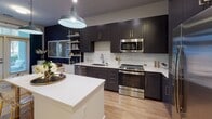 Located at 2100 South Blvd. in Charlotte's vibrant South End neighborhood, this single-floor residence blends contemporary design with thoughtful detail throughout. The open-concept kitchen is a standout, featuring dark shaker-style cabinetry with gold hardware, crisp white quartz countertops, a classic subway tile backsplash, and stainless steel appliances including a range, microwave, and dishwasher. A central island provides additional prep space and casual seating, while an industrial-style pendant light anchors the room. Light hardwood flooring flows through the main living areas, complemented by open shelving that adds functional style. The living area benefits from large windows dressed with deep green curtains, allowing generous natural light and views of the surrounding neighborhood. A sculptural mirror and fluted white sideboard contribute to the space's refined aesthetic. The bedroom offers a calm retreat, highlighted by a bold teal accent wall adorned with large-scale macramé wall art, plush carpeting, a ceiling fan, and ample natural light. The full bathroom is finished with dark large-format tile surrounding a soaking tub and shower combination, a dark-cabineted vanity with white countertop, and a broad frameless mirror, delivering a clean, modern feel throughout.