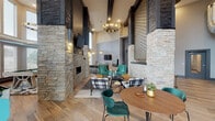 Located at 1330 Kelly Johnson Blvd in Colorado Springs, CO, this single-floor amenity space delivers a striking blend of industrial and contemporary design. Soaring double-height ceilings create an expansive, airy atmosphere throughout, while floor-to-ceiling stacked stone columns serve as dramatic architectural focal points that anchor the open-concept layout. Warm hardwood flooring flows seamlessly across the space, contrasting beautifully with the natural stone and dark wood beam accents overhead.

The central lounge area is defined by bold, eclectic furnishings — teal leather armchairs, plaid sofas, mustard ottomans, and a geometric patterned area rug — all centered around a sleek stone fireplace with a linear hearth. A decorative lattice-panel feature wall adds textural depth and visual interest. Stylish pendant and chandelier lighting enhances the ambiance throughout.

A quieter seating nook offers a pair of modern rounded chairs flanking a secondary fireplace, ideal for more intimate conversation. The game area is generously sized, featuring a pool table set beneath a dramatic wall of tall windows that flood the room with natural light, alongside a vintage arcade game for added entertainment. A wet bar and additional dining and seating areas complete this well-appointed communal space, designed for both socializing and relaxation.