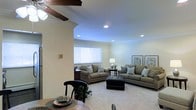 This single-floor residence offers a thoughtfully designed layout centered around a spacious, open-concept living area. The living room is a standout feature, showcasing recessed lighting that casts a warm, even glow across the room, complemented by a ceiling fan that adds both function and a classic touch. Crown molding frames the walls and ceiling, lending an air of refined elegance throughout the space. Plush wall-to-wall carpeting in a neutral tone creates a comfortable and inviting atmosphere, while large windows dressed with white horizontal blinds allow for generous natural light and a sense of openness. The living area comfortably accommodates multiple seating arrangements, with ample room for sofas, accent chairs, and a central ottoman, all anchored by tasteful framed artwork on the walls. A structural column with decorative molding adds architectural character and subtly defines the transition between the living and dining areas. The dining space, visible in the foreground, features a warm-toned table and classic dark wood chairs, creating a welcoming setting for meals. Overall, the home presents a clean, neutral palette with traditional architectural details that combine to create a warm, livable, and well-balanced interior.