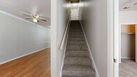 This two-story home offers a practical and spacious floor plan with five bedrooms, two full bathrooms, and generous living areas spread across two levels. The main floor accounts for the majority of the living space and features warm hardwood-style flooring in the living area, accented by crown molding and a ceiling fan. The galley-style kitchen is outfitted with dark cabinetry, white countertops, black appliances including a range, refrigerator, and dishwasher, and light tile flooring. A door with a window panel leads directly to the exterior, allowing natural light into the kitchen. The bathrooms feature tile flooring, dark vanity cabinetry, white countertops, and a tub-shower combination. The bedrooms are carpeted and include ceiling fans and windows that bring in natural light. Four closets throughout the home provide ample storage. A dedicated laundry room includes washer and dryer hookups, a water heater, and tile flooring. Carpeted staircases connect the two floors, with clean white railings and handrails. The upper level provides additional bedroom space. Outside, a fenced backyard with mature trees offers a private outdoor area, complemented by a central air conditioning unit.