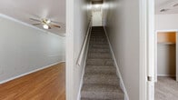 This two-story home offers a practical and spacious floor plan with five bedrooms, two full bathrooms, and generous living areas spread across two levels. The main floor accounts for the majority of the living space and features warm hardwood-style flooring in the living area, accented by crown molding and a ceiling fan. The galley-style kitchen is outfitted with dark cabinetry, white countertops, black appliances including a range, refrigerator, and dishwasher, and light tile flooring. A door with a window panel leads directly to the exterior, allowing natural light into the kitchen. The bathrooms feature tile flooring, dark vanity cabinetry, white countertops, and a tub-shower combination. The bedrooms are carpeted and include ceiling fans and windows that bring in natural light. Four closets throughout the home provide ample storage. A dedicated laundry room includes washer and dryer hookups, a water heater, and tile flooring. Carpeted staircases connect the two floors, with clean white railings and handrails. The upper level provides additional bedroom space. Outside, a fenced backyard with mature trees offers a private outdoor area, complemented by a central air conditioning unit.