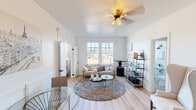 This single-floor unit offers a thoughtfully designed layout that balances comfort and style throughout its key living spaces. The living room serves as the heart of the unit, featuring light hardwood flooring, crisp white walls, and an abundance of natural light streaming through large gridded windows. A ceiling fan with warm-toned lighting anchors the room, which is complemented by a plush area rug, a cushioned sofa, a glass coffee table, and an open metal shelving unit that adds an industrial accent. A classic wingback chair provides an additional seating nook, while a small dining area with a glass-top table rounds out the space.

The bedroom is an intimate and serene retreat, highlighted by a window that frames outdoor views and allows natural light to fill the room. White walls and hardwood flooring continue the cohesive aesthetic established throughout the unit.

The kitchen is compact yet functional, outfitted with a gas range, a stainless steel sink, a refrigerator, and a subway tile backsplash. A wall-mounted range hood and open wooden shelving add both practicality and character to the space.

The bathroom is a standout feature, showcasing floor-to-ceiling white subway tile surrounding the tub and shower, accented by matte black fixtures. A window above the tub fills the room with natural light while offering a pleasant outdoor view, completing the clean, modern aesthetic of this well-appointed unit.