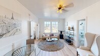 This single-floor unit offers a thoughtfully designed layout that balances comfort and style throughout its key living spaces. The living room serves as the heart of the unit, featuring light hardwood flooring, crisp white walls, and an abundance of natural light streaming through large gridded windows. A ceiling fan with warm-toned lighting anchors the room, which is complemented by a plush area rug, a cushioned sofa, a glass coffee table, and an open metal shelving unit that adds an industrial accent. A classic wingback chair provides an additional seating nook, while a small dining area with a glass-top table rounds out the space.

The bedroom is an intimate and serene retreat, highlighted by a window that frames outdoor views and allows natural light to fill the room. White walls and hardwood flooring continue the cohesive aesthetic established throughout the unit.

The kitchen is compact yet functional, outfitted with a gas range, a stainless steel sink, a refrigerator, and a subway tile backsplash. A wall-mounted range hood and open wooden shelving add both practicality and character to the space.

The bathroom is a standout feature, showcasing floor-to-ceiling white subway tile surrounding the tub and shower, accented by matte black fixtures. A window above the tub fills the room with natural light while offering a pleasant outdoor view, completing the clean, modern aesthetic of this well-appointed unit.