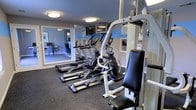 This fitness center offers a well-organized workout space designed to meet a variety of exercise needs. The room features durable rubber flooring, which provides both safety and comfort during workouts. Several elliptical trainers are lined up along one side, offering ample cardio options for users. A multi-station cable and weight machine anchors the strength training area, complemented by an adjustable bench seat for a range of exercises. A wall-mounted television is positioned for easy viewing during cardio sessions, adding convenience to longer workouts. The walls are painted in a neutral tone accented by a blue horizontal stripe, giving the space a clean, structured aesthetic. Flush-mount ceiling lights provide consistent, even illumination throughout the room. A large pass-through window connects the fitness center to an adjacent lounge or common area, adding a sense of openness and visual connectivity. Informational notices and safety guidelines are posted on the walls, reflecting attention to user awareness. The overall layout is efficient and functional, making effective use of the available space while keeping equipment accessible and organized.