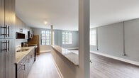 This single-floor unit is designed with a clean, modern aesthetic throughout. The open-concept layout connects the kitchen directly to the living area, creating a seamless and functional flow. The kitchen features warm wood-toned cabinetry, a granite-topped breakfast bar, subway tile backsplash, and stainless steel appliances including a refrigerator, range, and microwave. Track lighting above the kitchen adds focused illumination to the workspace.

The living area is generously proportioned, benefiting from multiple windows dressed with vertical blinds that allow ample natural light to fill the space. Neutral gray walls complement the light wood-look vinyl flooring found throughout the main living zones. A ductless mini-split system provides climate control in both the living area and the bedroom, offering efficient and independent temperature management.

The bedroom is a well-sized, quiet retreat with its own mini-split unit, a window for natural light, and durable wood-look flooring. The bathroom is finished with tile flooring, white wall tile, a pedestal-style sink, overhead cabinetry for storage, and a glass-enclosed walk-in shower with brushed nickel fixtures, delivering a crisp and functional design.