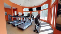 Located at 3301 Glenn McConnell Parkway in Charleston, South Carolina, this fitness center offers a well-equipped, single-floor workout facility designed to energize and motivate. The space is immediately distinguished by its bold orange walls, which create a vibrant and high-energy atmosphere throughout. Soaring vaulted ceilings with recessed lighting and ceiling fans add a sense of openness and comfort to the environment.

The cardio area features a curved, windowed bay lined with large arched windows that flood the room with natural light and offer views of the surrounding wooded landscape. Multiple treadmills, elliptical trainers, and stationary bikes are arranged to face wall-mounted flat-screen televisions, providing an engaging workout experience. The flooring throughout is durable carpeting suited for fitness use.

A separate strength training area accommodates a variety of equipment, including a cable machine, weight benches, an adjustable incline bench, a dumbbell rack, and additional resistance machines. Full-length mirrors along the walls enhance the sense of space and allow for proper form checks during exercise. A dedicated exit door with sidelights provides direct outdoor access, adding convenience and ventilation options. Together, these spaces form a comprehensive, well-lit fitness facility.
