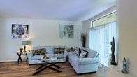 This single-floor space offers a comfortable and thoughtfully arranged living environment with a clean, neutral aesthetic. The living room serves as the heart of the space, featuring warm hardwood flooring that adds a sense of warmth and continuity throughout. Soft, light-toned walls create an airy atmosphere that is both calming and inviting.

The living area is well-appointed with plush seating arranged around a glass-topped coffee table, offering a functional yet stylish layout ideal for relaxing or entertaining. Decorative accents, including patterned throw pillows and a botanical wall piece, add subtle character without overwhelming the space.

A standout feature of the living room is the large sliding glass door fitted with vertical blinds, which allows generous natural light to fill the room while providing privacy and light control. The door offers a seamless transition between the interior and an outdoor area beyond.

Overall, the space presents a bright, well-maintained interior with a cohesive design that balances comfort and practicality. The combination of natural light, warm flooring, and understated décor gives the space a welcoming ambiance suited to everyday living.