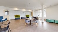 Located at 6834 Milwaukee St in Madison, Wisconsin, this single-floor office space offers a well-appointed, open-plan layout designed to accommodate a variety of work and meeting needs. The interior is finished with large-format neutral tile flooring throughout, white walls, and smooth ceilings with recessed lighting complemented by distinctive circular pendant fixtures, creating a bright and professional atmosphere.

The space is organized into several functional zones. A central round table with upholstered grey chairs serves as a collaborative hub, ideal for group discussions or informal meetings. Multiple individual workstations and desks are positioned around the perimeter, offering dedicated areas for focused work. A comfortable lounge corner features bold blue accent chairs alongside a vibrant abstract wall art piece, adding a creative touch to the environment. A teal modular seating unit near the entrance provides an additional casual gathering spot.

Large windows on multiple walls flood the space with natural light and offer views of the surrounding exterior. Structural columns are integrated throughout the open floor plan. Decorative tall grass planters in navy pots add a natural, organic element to the space. A printer station and additional worktables round out the practical amenities. Direct access to the exterior is provided via a glass-panel door, enhancing connectivity and ease of access.