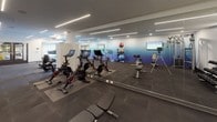 This fitness center offers a thoughtfully designed space for a comprehensive workout experience. The room features a striking ombre accent wall that transitions from deep navy blue to lighter tones, creating an energizing atmosphere complemented by neutral grey walls and ceiling. Overhead, a combination of recessed lighting and linear LED strip fixtures provides consistent, bright illumination throughout the space.

The flooring consists of durable dark rubber tiles, dividing the room into functional workout zones. The cardio section includes elliptical machines, stationary spin bikes equipped with digital screens, a rowing machine, and a stair climber. A dedicated strength training area features a cable machine, adjustable benches, free weights, and a back extension bench. Stability balls of varying sizes are neatly stored on a vertical rack, adding versatility for core training.

Multiple wall-mounted flat-screen televisions are positioned throughout the room, allowing users to stay entertained during workouts. A large floor-to-ceiling mirror along one wall expands the sense of space. Entry is through glass-paneled double doors, which connect the fitness center to adjacent community areas, ensuring convenient access for all users.