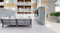 Located at 2840-2850 Carlow Rd in Victoria, BC, this outdoor amenity courtyard is a well-appointed communal space nestled between the multi-storey residential building and its surrounding structures. The courtyard is finished with large-format porcelain tiles throughout, creating a clean, contemporary aesthetic that complements the building's modern exterior of grey paneling and warm wood-toned cladding.

The space is organized into several distinct activity and relaxation zones. A dedicated bocce ball court with a wood-framed turf surface provides a fun recreational focal point, while a lounge area nearby features comfortable outdoor sofas and armchairs arranged around a built-in fireplace unit — ideal for year-round enjoyment. String lights are strung overhead in portions of the courtyard, adding an inviting ambiance during evening hours.

Generous raised planters filled with lush tropical palms and greenery introduce natural elements and visual privacy, with integrated wooden benches along their bases offering additional casual seating. An outdoor pergola structure anchors one end of the courtyard, further defining the space. The open-air design allows for abundant natural light, and the surrounding building architecture provides a sense of enclosure and intimacy. This thoughtfully designed amenity space offers residents a versatile outdoor retreat for both recreation and relaxation.
