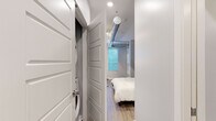 This smartly designed one-bedroom, one-bathroom unit makes the most of its compact footprint through thoughtful layout and modern finishes. The open floor plan integrates the living and sleeping areas seamlessly, anchored by a sleek Murphy bed with a dark-framed surround that folds away to maximize usable floor space. Light wood-look flooring runs throughout, lending warmth and visual continuity across all areas.

The kitchenette is neatly built into one wall of the main room and features white shaker cabinetry, a full suite of black appliances including a refrigerator, range, microwave, and dishwasher, plus a single-basin sink. A small bar-height counter area provides a convenient spot for dining or workspace.

The bedroom area benefits from natural light streaming through horizontal-slatted windows that overlook greenery outside. An exposed metallic ventilation duct runs diagonally across the ceiling, adding an industrial design accent to the otherwise clean, contemporary aesthetic with soft gray walls.

The hallway connects the main living space to the bathroom, which is finished with light wood-look flooring, a white vanity with ample cabinet storage, a large mirror with overhead lighting, and a glass-door shower enclosure. A walk-in closet is visible through the bathroom, offering additional storage.

Overall, this unit delivers a practical and polished living experience with smart space-saving solutions throughout.