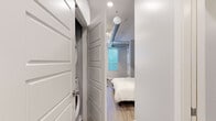 This smartly designed one-bedroom, one-bathroom unit makes the most of its compact footprint through thoughtful layout and modern finishes. The open floor plan integrates the living and sleeping areas seamlessly, anchored by a sleek Murphy bed with a dark-framed surround that folds away to maximize usable floor space. Light wood-look flooring runs throughout, lending warmth and visual continuity across all areas.

The kitchenette is neatly built into one wall of the main room and features white shaker cabinetry, a full suite of black appliances including a refrigerator, range, microwave, and dishwasher, plus a single-basin sink. A small bar-height counter area provides a convenient spot for dining or workspace.

The bedroom area benefits from natural light streaming through horizontal-slatted windows that overlook greenery outside. An exposed metallic ventilation duct runs diagonally across the ceiling, adding an industrial design accent to the otherwise clean, contemporary aesthetic with soft gray walls.

The hallway connects the main living space to the bathroom, which is finished with light wood-look flooring, a white vanity with ample cabinet storage, a large mirror with overhead lighting, and a glass-door shower enclosure. A walk-in closet is visible through the bathroom, offering additional storage.

Overall, this unit delivers a practical and polished living experience with smart space-saving solutions throughout.