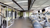 This fitness center offers a comprehensive workout environment designed to accommodate a range of exercise needs. The space features an industrial-inspired aesthetic, with an exposed ceiling showcasing structural beams, visible ductwork, and coffered panels illuminated by rows of linear fluorescent lighting, lending the room an open, loft-like character.

Along one wall, floor-to-ceiling windows provide abundant natural light and views of an adjacent outdoor green space and surrounding buildings, creating an airy atmosphere during workouts. The cardio zone lines this windowed wall and features a row of Precor treadmills, elliptical trainers, and upright stationary bikes, each equipped with individual mounted flat-screen televisions. Large mirrors along the interior walls visually expand the space and allow users to monitor their form throughout their sessions.

The strength training area is positioned on the opposite side of the floor and includes a multi-functional cable machine system, an adjustable weight bench, and free weights including kettlebells. The durable rubber flooring throughout provides both safety and practicality. Overall, the fitness center presents a functional and well-organized environment suited for both cardio and strength training.