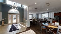 This single-floor community clubhouse is thoughtfully designed to offer residents a range of amenities in a welcoming and comfortable environment. The entrance opens into a spacious common area distinguished by elegant white columns, soaring ceilings with clerestory windows, and large arched glass doors that flood the space with abundant natural light. The soft blue-gray walls and patterned carpeting lend the area a calm, cohesive aesthetic throughout.

The dining and lounge area features a mix of round tables with cross-back chairs and bar-height seating along a granite-topped counter, offering flexible arrangements for casual dining or social gatherings. Crown molding and recessed lighting add a refined architectural touch. A cozy sitting nook with a classic white-mantled fireplace, wall-mounted television, and leather armchairs provides an inviting retreat, bathed in natural light from large windows with exterior views.

The kitchen is equipped with rich cherry wood cabinetry, granite countertops, and a full suite of stainless steel appliances including a range, microwave, refrigerator, and dishwasher — well-suited for resident use or event catering.

The fitness center is generously sized and outfitted with multiple treadmills, elliptical trainers, stationary bikes, and cable strength machines. Oversized windows provide views of the surrounding community, keeping the space bright and energizing throughout the day.