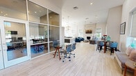 Located at 901 W Little Creek Rd in Norfolk, Virginia, this single-floor community clubhouse offers a thoughtfully designed, open-plan layout with a welcoming and modern aesthetic. Light hardwood-style flooring flows throughout, complementing the soaring ceilings and abundant natural light streaming through large, Roman shade-dressed windows.

The expansive main area serves multiple functions, anchored by a striking stacked-stone fireplace with a wall-mounted television, framed by plush blue sofas and casual seating — an inviting lounge ideal for relaxing. A pool table adds a recreational dimension to the space, alongside a classic popcorn cart that lends a playful touch.

The kitchenette is a standout feature, centered around a large quartz-topped island with upholstered seating. Elegant gold geometric pendant lights hang above, while the backsplash wall features a bold striped tile pattern. Stainless steel appliances, ample cabinetry, and a full-size refrigerator complete the well-equipped prep area.

A glass-walled private conference room offers a dedicated workspace with a central table, task seating, computers, and a sculptural pendant light. A cozy seating nook with a round glass table basks in natural light from corner windows with outdoor views. The space proudly displays a "We Support Our Troops" banner, reflecting its community-oriented character.