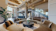 This expansive single-floor communal space is defined by its dramatic double-height ceilings and open-plan layout, creating an airy, grand atmosphere throughout. The design blends contemporary and transitional aesthetics, combining warm wood accents on coffered ceiling insets with cool gray structural columns and polished large-format tile flooring in neutral tones.

The central lounge area anchors the space with curved, velvet sectional sofas arranged around a dark, cylindrical coffee table, offering a refined seating environment beneath layered modern chandelier lighting. A sleek staircase with dark treads, integrated step lighting, and a glass-panel railing serves as a prominent architectural focal point.

Adjacent to the lounge, a fully appointed bar area features an extended dark stone countertop, backlit tile walls, a wall-mounted television, and rows of contemporary bar stools. Nearby dining zones are furnished with dark stone-top tables and mid-century-inspired wood chairs.

Floor-to-ceiling window walls flood the space with abundant natural light while providing expansive outdoor views. Additional seating nooks with warm tile floors and eclectic furnishings provide intimate retreat spaces. Tropical potted plants are positioned throughout, enhancing the welcoming, upscale ambiance of the space.