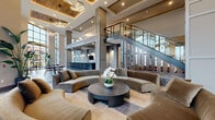 This expansive single-floor communal space is defined by its dramatic double-height ceilings and open-plan layout, creating an airy, grand atmosphere throughout. The design blends contemporary and transitional aesthetics, combining warm wood accents on coffered ceiling insets with cool gray structural columns and polished large-format tile flooring in neutral tones.

The central lounge area anchors the space with curved, velvet sectional sofas arranged around a dark, cylindrical coffee table, offering a refined seating environment beneath layered modern chandelier lighting. A sleek staircase with dark treads, integrated step lighting, and a glass-panel railing serves as a prominent architectural focal point.

Adjacent to the lounge, a fully appointed bar area features an extended dark stone countertop, backlit tile walls, a wall-mounted television, and rows of contemporary bar stools. Nearby dining zones are furnished with dark stone-top tables and mid-century-inspired wood chairs.

Floor-to-ceiling window walls flood the space with abundant natural light while providing expansive outdoor views. Additional seating nooks with warm tile floors and eclectic furnishings provide intimate retreat spaces. Tropical potted plants are positioned throughout, enhancing the welcoming, upscale ambiance of the space.