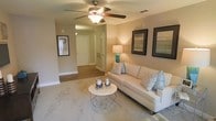 This single-story space centers around a comfortable living area finished in soft, neutral beige tones that create a calm and inviting atmosphere. The room features plush carpet underfoot, providing a cozy feel, while a ceiling fan with an integrated light fixture serves as the functional focal point overhead. The walls are adorned with framed artwork, contributing a polished, curated aesthetic to the space.

A cream-toned sofa anchors the seating area, complemented by patterned accent pillows in muted blues and grays. A geometric wire-frame coffee table with a round marble-style top adds a contemporary touch to the otherwise transitional design. A teal table lamp introduces a subtle pop of color and warm ambient lighting to the corner of the room.

The living area opens directly into a hallway, where hardwood-style flooring transitions from the carpeted living space, adding visual contrast. The hallway leads to additional rooms, hinting at a practical and well-connected layout. White interior doors and clean white baseboards throughout the space enhance the bright, tidy feel. Overall, the space presents a cohesive, neutral palette with thoughtful design details throughout.