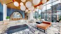 Located on Colshire Drive in McLean, Virginia, this sophisticated single-floor lounge and amenity space offers a striking architectural experience defined by grandeur and thoughtful design. The space is anchored by dramatically tall ceilings adorned with a cascading cluster of oversized white globe pendant lights, creating a warm, inviting ambiance that draws the eye upward.

Floor-to-ceiling glass walls line the perimeter, flooding the interior with abundant natural light and offering sweeping views of the surrounding McLean skyline. The open lounge area features generously sized neutral sofas, leather chaise lounges, and sculptural coffee tables arranged atop a bold abstract area rug, blending comfort with contemporary aesthetics.

A standout bar and kitchenette zone showcases a bold arched architectural feature framed in deep blue tones with an antiqued mirror backdrop, complemented by a granite-topped island with wood-framed barstools. The flooring transitions artfully between herringbone hardwood and geometric inlaid tile patterns, adding visual interest throughout.

Large sliding glass doors open directly onto an outdoor terrace featuring manicured turf, patio seating, and open-air entertaining areas, seamlessly connecting interior and exterior living. The overall atmosphere is modern, refined, and resort-inspired.