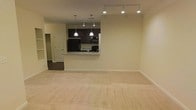 This single-floor space features an open-concept layout that seamlessly connects the living area to the kitchen. The living area is finished with plush, neutral-toned carpeting that adds warmth and comfort underfoot, while the surrounding walls are painted in a soft, light cream hue that enhances the sense of openness. Track lighting runs along the ceiling, providing flexible and even illumination throughout the space.

The kitchen is positioned at the rear and is accessible via a peninsula-style pass-through counter topped with light granite or stone-look surfaces, offering both a visual divider and a casual dining or prep area. Dark espresso-toned cabinetry lines the kitchen walls, creating a striking contrast against the lighter surroundings. Two pendant lights hang above the counter, adding a focused decorative touch. The kitchen is equipped with black appliances, including a refrigerator, range, and microwave, contributing to a cohesive, modern aesthetic.

A doorway visible to the left leads to an additional room, suggesting further functional space beyond the main living area. Overall, the space maintains a clean, neutral, and move-in-ready ambiance suitable for a variety of furnishing styles.