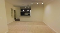 This single-floor space features an open-concept layout that seamlessly connects the living area to the kitchen. The living area is finished with plush, neutral-toned carpeting that adds warmth and comfort underfoot, while the surrounding walls are painted in a soft, light cream hue that enhances the sense of openness. Track lighting runs along the ceiling, providing flexible and even illumination throughout the space.

The kitchen is positioned at the rear and is accessible via a peninsula-style pass-through counter topped with light granite or stone-look surfaces, offering both a visual divider and a casual dining or prep area. Dark espresso-toned cabinetry lines the kitchen walls, creating a striking contrast against the lighter surroundings. Two pendant lights hang above the counter, adding a focused decorative touch. The kitchen is equipped with black appliances, including a refrigerator, range, and microwave, contributing to a cohesive, modern aesthetic.

A doorway visible to the left leads to an additional room, suggesting further functional space beyond the main living area. Overall, the space maintains a clean, neutral, and move-in-ready ambiance suitable for a variety of furnishing styles.