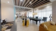This single-floor amenity space is thoughtfully designed to offer a dynamic and welcoming social environment. The open-plan layout allows distinct activity zones to flow seamlessly into one another, creating a sense of expansiveness throughout.

At the heart of the space sits a full-size pool table, surrounded by stylish bar-height stools in bold yellow and black — a striking focal point that anchors the entertainment area. Overhead, dramatic dark exposed wood beams contrast beautifully against the white ceiling, adding warmth and architectural character to the room.

Large floor-to-ceiling windows along one wall flood the interior with natural light, softened by sheer white drape panels. Pendant lighting with woven shades hangs gracefully above, complementing the modern yet relaxed aesthetic.

Comfortable lounge seating in teal and mustard tones is arranged throughout, providing ample space for casual socializing and relaxation. A accent wall in aqua-blue adds a pop of color that enlivens the overall palette.

Toward the rear, a fully equipped kitchenette with stainless steel appliances and bar-style seating offers a functional space for gatherings. Large-format tile flooring ties the entire space together, delivering a clean, contemporary finish that is both practical and visually appealing.