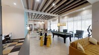 This single-floor amenity space is thoughtfully designed to offer a dynamic and welcoming social environment. The open-plan layout allows distinct activity zones to flow seamlessly into one another, creating a sense of expansiveness throughout.

At the heart of the space sits a full-size pool table, surrounded by stylish bar-height stools in bold yellow and black — a striking focal point that anchors the entertainment area. Overhead, dramatic dark exposed wood beams contrast beautifully against the white ceiling, adding warmth and architectural character to the room.

Large floor-to-ceiling windows along one wall flood the interior with natural light, softened by sheer white drape panels. Pendant lighting with woven shades hangs gracefully above, complementing the modern yet relaxed aesthetic.

Comfortable lounge seating in teal and mustard tones is arranged throughout, providing ample space for casual socializing and relaxation. A accent wall in aqua-blue adds a pop of color that enlivens the overall palette.

Toward the rear, a fully equipped kitchenette with stainless steel appliances and bar-style seating offers a functional space for gatherings. Large-format tile flooring ties the entire space together, delivering a clean, contemporary finish that is both practical and visually appealing.