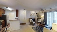 This single-floor residence offers a warm and inviting open-concept layout that seamlessly connects the living area and kitchen. The living room serves as the focal point of the space, featuring a tiled fireplace with a classic white mantle, built-in shelving for display, and a ceiling fan overhead. A sliding glass door dressed with vertical blinds and draping curtain panels fills the room with natural light while providing outdoor access. The textured ceiling and neutral-toned carpet throughout contribute to the space's cozy ambiance.

Seating is thoughtfully arranged around a central wooden coffee table, with a plush sofa, wicker accent chairs, and layered rugs anchoring the room. Warm lamp lighting complements the natural light, creating an inviting atmosphere throughout the day and evening.

The kitchen is positioned adjacent to the living area, separated by a raised breakfast bar with a granite-style countertop and dark wood cabinetry above. A front entry with a solid wood door and a tiled foyer provides a clear transition into the main living space, keeping the layout practical and well-organized.