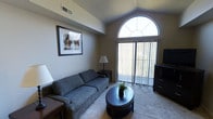 This single-floor space offers a well-appointed living area that immediately draws the eye upward with its vaulted ceilings, adding a sense of openness and volume to the room. A striking arched window serves as the architectural centerpiece, flooding the space with natural light and creating an elegant focal point above the sliding glass door, which opens to an outdoor balcony with views of surrounding greenery. The living area is finished with neutral-toned carpeting and warm beige walls, providing a versatile backdrop for the furnishings. The room comfortably accommodates a full-sized sofa, a rounded dark-wood coffee table, and a media console, creating a functional and cohesive layout. Vertical blinds on the sliding door allow for flexible light and privacy control. A framed piece of wall art adds a touch of character to the space. Overhead ventilation is integrated discreetly into the ceiling, ensuring comfort throughout. The overall ambiance is calm and inviting, combining architectural details like the arched transom window with practical amenities to create a livable, light-filled environment.