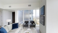 This single-floor residence exemplifies contemporary urban living through its clean architectural lines, neutral palette, and thoughtfully curated design. The living area serves as the centerpiece of the space, anchored by expansive floor-to-ceiling windows that frame sweeping high-rise city and treetop views, flooding the interior with abundant natural light. The crisp white walls and light-toned tile flooring create a bright, airy atmosphere that enhances the sense of openness throughout. Elegant velvet accent chairs in charcoal grey are paired with a plush white sofa adorned with deep navy cushions, striking a refined balance between comfort and sophistication. A sleek wall-mounted display panel adds a modern, minimalist touch to the main wall. Frosted glass privacy panels complement the clear glazing, offering a layered approach to light diffusion without sacrificing the connection to the outdoors. Recessed ceiling lighting and strategically placed ventilation maintain an unobtrusive, polished aesthetic. The overall ambiance is one of understated luxury — calm, curated, and elevated — making the space well-suited for both quiet relaxation and refined entertaining.