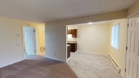 This single-floor space offers a clean, open layout designed for comfortable everyday living. The main living area is finished with plush carpeting in warm neutral tones, creating a cozy atmosphere that transitions seamlessly into the kitchen area via an open pass-through design. The kitchen nook features dark wood cabinetry with ample upper and lower storage, a light-toned countertop, and light wood-look tile flooring that adds a modern, easy-to-maintain touch. The open connection between the living and kitchen spaces enhances the sense of flow and openness throughout the unit. Walls are painted in soft, warm neutral tones that complement the lighter flooring and trim, lending the space a bright and inviting feel. Recessed lighting in the kitchen area ensures the workspace is well illuminated. A bathroom is accessible from the main living area, featuring tile flooring, a toilet, and clean white fixtures, with a door offering full privacy. White panel doors and crisp white baseboards provide classic finishing details throughout. The textured ceiling adds a traditional architectural element. Overall, the space presents a functional and well-maintained interior with a cohesive, neutral aesthetic suitable for a variety of uses.