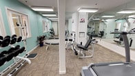 This fitness center offers a comprehensive workout environment designed to accommodate a wide range of exercise routines. The space is finished with soft teal-painted walls and neutral, textured carpet flooring, creating a clean and energizing atmosphere. Recessed panel lighting illuminates the room evenly throughout, ensuring excellent visibility across all workout areas.

The strength training section is anchored by multiple Cybex cable and resistance machines, providing users with professional-grade equipment for targeted muscle work. A dedicated free weights area features a multi-tier dumbbell rack stocked with a full range of weights, from light pink and blue dumbbells to heavy black sets, alongside a flat weight bench for added versatility. Potted plants are thoughtfully placed throughout, adding a touch of greenery to the space.

The cardio zone includes a treadmill and an elliptical trainer, offering options for both high- and low-impact aerobic workouts. Full-length mirrors line the walls, visually expanding the space and allowing users to monitor their form during exercise. A wire rack near the free weights area holds fresh towels for convenience. The fitness center is accessed through a dedicated entry door and is clearly marked with emergency exit signage, reflecting a commitment to safety and accessibility.