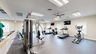 This fitness center is designed to accommodate a comprehensive range of workout routines within a spacious, open-plan single-floor layout. The room features durable wood-look flooring throughout, providing a clean and practical surface suited to high-traffic exercise use. Recessed flush-panel lighting is distributed evenly across the ceiling, supplemented by multiple ceiling fans that promote consistent airflow and comfort during workouts. Natural light enters through several windows fitted with white blinds along the perimeter walls, contributing to a bright and welcoming atmosphere. Neutral beige walls are accented with decorative artwork, adding a polished, inviting finish to the space.

The cardio section is well-stocked with multiple treadmills, upright and recumbent stationary bikes, and an elliptical trainer — several from the Spirit brand — positioned to allow ample space between each machine. Wall-mounted flat-screen televisions are positioned for easy viewing from both the cardio and strength training areas. A multi-station cable machine anchors the strength training zone, while a full rack of dumbbells offers a wide range of free weight options. Functional training accessories include stability balls, resistance bands, and exercise mats organized on a dedicated storage rack. A mirror wall enhances the sense of depth. The facility connects to an adjacent hallway, suggesting it is part of a larger shared amenity complex.