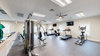This fitness center is designed to accommodate a comprehensive range of workout routines within a spacious, open-plan single-floor layout. The room features durable wood-look flooring throughout, providing a clean and practical surface suited to high-traffic exercise use. Recessed flush-panel lighting is distributed evenly across the ceiling, supplemented by multiple ceiling fans that promote consistent airflow and comfort during workouts. Natural light enters through several windows fitted with white blinds along the perimeter walls, contributing to a bright and welcoming atmosphere. Neutral beige walls are accented with decorative artwork, adding a polished, inviting finish to the space.

The cardio section is well-stocked with multiple treadmills, upright and recumbent stationary bikes, and an elliptical trainer — several from the Spirit brand — positioned to allow ample space between each machine. Wall-mounted flat-screen televisions are positioned for easy viewing from both the cardio and strength training areas. A multi-station cable machine anchors the strength training zone, while a full rack of dumbbells offers a wide range of free weight options. Functional training accessories include stability balls, resistance bands, and exercise mats organized on a dedicated storage rack. A mirror wall enhances the sense of depth. The facility connects to an adjacent hallway, suggesting it is part of a larger shared amenity complex.