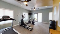 This single-level fitness center is designed to provide a comfortable and functional workout environment. The space is finished with neutral patterned carpeting and soft sage-green walls that create a calm, focused atmosphere throughout. Dual ceiling fans with integrated lighting ensure consistent airflow and comfort during exercise sessions, complemented by recessed lighting for even illumination.

The cardio area is well-equipped, featuring a treadmill, elliptical trainer, and a recumbent stationary bike, giving users a variety of options for cardiovascular workouts. A wall-mounted flat-screen television is positioned for easy viewing during exercise. The strength training area includes a weight bench and a fully stocked dumbbell rack, accommodating a range of resistance training exercises.

A striking yellow accent wall adds a bold visual element to the strength training section, providing contrast against the otherwise neutral palette. Large windows fitted with roller shades allow natural light to filter in while maintaining privacy. A glass-panel door opens directly to an outdoor courtyard area, offering convenient access and additional ventilation. Free Wi-Fi is available throughout the space, and the overall layout is open and spacious, allowing users to move freely between equipment zones.