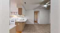 This single-story home is designed with a functional, open layout that connects the kitchen and living areas seamlessly. The kitchen is a standout feature, equipped with light wood cabinetry, white appliances including an electric range, dishwasher, and refrigerator, and a peninsula with a built-in sink that overlooks the living space. A vaulted ceiling with an angled soffit adds architectural interest, while a ceiling fan provides circulation throughout the open area. The kitchen transitions from hardwood-style flooring to carpeted living areas, creating a natural distinction between zones.

The living area is bright and open, with neutral walls and plush carpet underfoot, leading to a hallway that connects to the remaining rooms. The bedrooms are carpeted and feature vertical blinds over windows that allow natural light to filter in. The bathroom includes a combination tub and shower with sliding glass doors and hardwood-style flooring. A dedicated laundry area accommodates a full-size washer and dryer side by side, with an overhead shelf for added storage. Throughout the home, the neutral color palette and clean finishes create a light and functional environment.