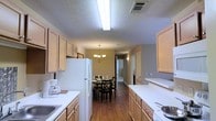 Located at 501 Fairways Dr in Vicksburg, Mississippi, this single-story residence offers a comfortable and functional living environment. The kitchen is a standout feature, designed in a galley-style layout with light wood cabinetry lining both sides, providing generous storage and counter space. White appliances, including a full-size refrigerator and range, complement the clean white countertops, while bright overhead fluorescent lighting ensures excellent visibility throughout the workspace. The kitchen flows seamlessly into a dedicated dining area, distinguished by rich hardwood flooring and an elegant chandelier that adds a warm, inviting ambiance to the space. The dining area comfortably accommodates a table and chairs set, making it well-suited for everyday meals or entertaining. Warm, neutral-toned walls throughout create a cohesive and welcoming atmosphere. A hallway beyond the dining area provides access to the remaining rooms of the home, suggesting a thoughtfully organized floor plan. The textured ceiling and consistent interior finishes reflect a practical yet comfortable residential design throughout this Vicksburg property.