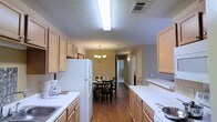 Located at 501 Fairways Dr in Vicksburg, Mississippi, this single-story residence offers a comfortable and functional living environment. The kitchen is a standout feature, designed in a galley-style layout with light wood cabinetry lining both sides, providing generous storage and counter space. White appliances, including a full-size refrigerator and range, complement the clean white countertops, while bright overhead fluorescent lighting ensures excellent visibility throughout the workspace. The kitchen flows seamlessly into a dedicated dining area, distinguished by rich hardwood flooring and an elegant chandelier that adds a warm, inviting ambiance to the space. The dining area comfortably accommodates a table and chairs set, making it well-suited for everyday meals or entertaining. Warm, neutral-toned walls throughout create a cohesive and welcoming atmosphere. A hallway beyond the dining area provides access to the remaining rooms of the home, suggesting a thoughtfully organized floor plan. The textured ceiling and consistent interior finishes reflect a practical yet comfortable residential design throughout this Vicksburg property.