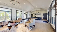Located at 1004 Bailes Ridge Ave in Fort Mill, SC, this single-floor community clubhouse offers a well-appointed gathering space defined by soaring vaulted ceilings and an open, airy layout. Large, dark-framed windows line the perimeter, flooding the interior with natural light and providing views of the surrounding landscape. The space is finished with large-format porcelain tile flooring, which contributes to the clean, contemporary aesthetic.

The lounge area is anchored by a white tile-surround fireplace flanked by dark, built-in shelving niches that provide both display space and architectural interest. A wall-mounted flat-screen television above the mantle and a circular pendant chandelier overhead complement the sophisticated design. Comfortable upholstered seating and area rugs define multiple conversation zones throughout.

A dedicated kitchenette and bar area features a large, rounded granite-topped island with barstool seating, pendant lighting, dark cabinetry, and a white subway tile backsplash. A coffee station, sink, and integrated appliances line the counter along the back wall, making the space well-suited for hosting events and gatherings. Glass entry doors provide easy access to the exterior, reinforcing the seamless connection between indoor and outdoor spaces.