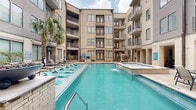Located at 3109 Douglas Avenue in Dallas, TX 75219, this outdoor amenity space serves as a vibrant communal hub designed for relaxation and social gathering. The centerpiece is a large, resort-style swimming pool with striking turquoise water, complemented by a raised spa with cascading water features. The pool deck is lined with teal chaise lounges and patio seating, creating an inviting sunbathing and outdoor dining environment, all framed by a multi-story building with private balconies overlooking the courtyard.

Adjacent to the pool is a covered outdoor living room that seamlessly blends indoor comfort with open-air access. This sheltered lounge area features exposed cream brick walls, polished concrete flooring, and ceiling fans that ensure comfortable airflow. The space is furnished with a curved sectional sofa in teal upholstery, accent artwork, and a wall-mounted flat-screen television. A built-in wet bar with dark cabinetry and granite countertops, along with a stainless steel refrigerator, provides convenient entertaining amenities. Tropical palm trees and lush greenery throughout the courtyard add a resort-like ambiance, making this a well-appointed outdoor retreat in the heart of Dallas.