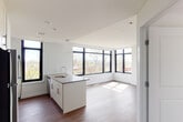 This contemporary single-floor residence is designed with a clean, modern aesthetic throughout. The open-concept living and kitchen area serves as the heart of the space, anchored by a large central island with a quartz-style countertop, an undermount sink, and ample cabinetry in a crisp white finish. The kitchen is fully equipped with stainless steel appliances, including a refrigerator, gas range, built-in microwave, and a green tile backsplash that adds a subtle accent. Floor-to-ceiling black-framed windows wrap around the living area, flooding the space with natural light and offering lush, tree-lined views that enhance the open, airy ambiance. Hardwood-style flooring runs throughout the main living areas, providing warmth and continuity. The bedroom is generously proportioned, featuring its own large black-framed window with treetop views and the same rich flooring. Two bathrooms serve the residence: one with a walk-in glass-enclosed shower and tile flooring, and a second full bathroom appointed with a soaking tub, handheld shower fixture, a wood-toned vanity with a mirror cabinet, and large-format tile floors. The overall design is cohesive and modern, emphasizing light, clean lines, and functional living.