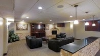 This single-floor space is thoughtfully designed as a cozy yet sophisticated entertainment and relaxation area. The room is anchored by a striking stacked-stone fireplace, which serves as a natural focal point and adds warmth and texture to the overall aesthetic. Adjacent to the fireplace, a rich dark-wood built-in media center provides ample storage with open shelving and cabinetry, seamlessly integrating the television into the room's design.

The seating arrangement features plush, dark quilted leather sofas and armchairs centered around a wooden coffee table, creating an inviting and cohesive lounge atmosphere. A pendant light fixture hangs above, complementing the recessed lighting embedded in the drop-tile ceiling, which provides even, layered illumination throughout the space.

The neutral-toned walls and carpeted flooring contribute to the room's understated elegance, while framed artwork adds a decorative touch. A stone-accented bar or accent table is partially visible in the foreground, hinting at additional entertaining features. Overall, the space balances functionality with comfort, making it well-suited for both casual gatherings and quiet relaxation.