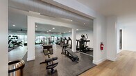 This single-floor fitness center is thoughtfully designed to support a full range of workout disciplines across several distinct zones. The space opens through a light-filled corridor finished with warm wood-plank flooring and clean white walls, providing a welcoming transition between areas. A glass-paneled studio room, clearly marked with signage, offers a dedicated space for group or individual classes.

The main workout floor is divided into well-defined sections. A cardio zone lines one wall with a row of commercial-grade treadmills and elliptical trainers, complemented by recumbent and upright cycling equipment nearby. A strength and functional training area features a comprehensive selection of free weights, adjustable benches, rowing machines, and cable machines, organized around rubber-matted flooring designed for high-impact use.

A separate strength machine room, flooded with natural light through large floor-to-ceiling windows overlooking a landscaped outdoor area, houses multiple selectorized resistance machines alongside dedicated stretching and mat space. Ceiling fans and exposed ductwork contribute to efficient ventilation throughout, while recessed lighting and arched windows add architectural character. The overall ambiance is modern, clean, and purposefully functional, offering members a comprehensive and comfortable environment for fitness.