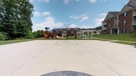 This outdoor recreational space in Jeffer offers a well-appointed community amenity area designed to serve residents of all ages. At the center of the space is a full-sized concrete basketball court, complete with a regulation backboard and hoop, providing a dedicated area for active play and sport. The smooth, expansive court surface is well-maintained and offers ample room for gameplay.

Adjacent to the court is a vibrant children's playground, featuring colorful play structures including slides and climbing elements, making it an engaging destination for younger residents. A covered pergola with decorative columns sits nearby, offering a shaded gathering spot ideal for relaxing or socializing outdoors.

The surrounding landscape is lush and green, with mature trees lining the perimeter and well-kept grassy areas framing the amenities. The backdrop includes multi-story brick residential buildings that contribute to the cohesive neighborhood character of the community. The open-sky setting allows for abundant natural light throughout the day, creating a welcoming and energetic outdoor environment. Overall, this recreational area presents a thoughtfully designed communal space that encourages outdoor activity and community engagement.